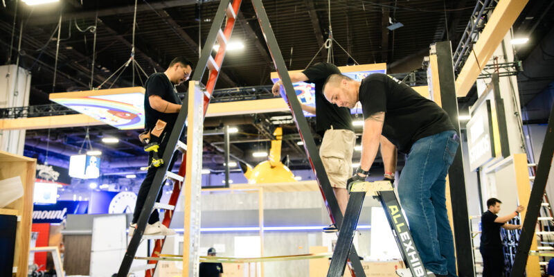 Crew member adjusting booth frame from ladder during setup.