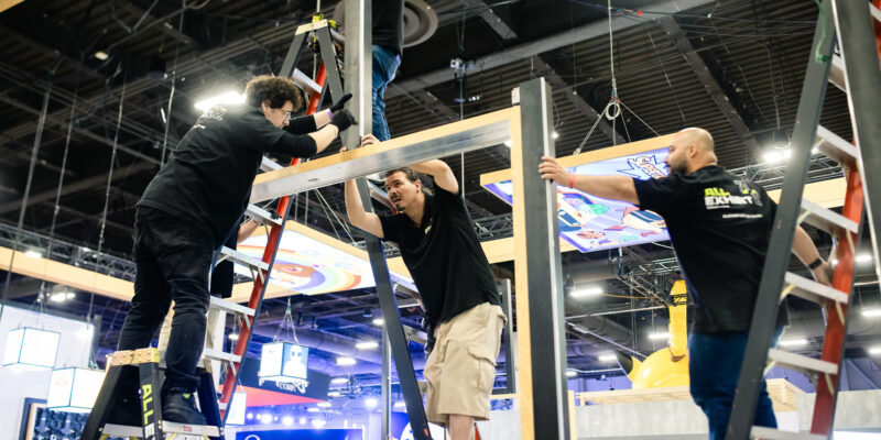 Workers on ladder installing booth frame beams at trade show.
