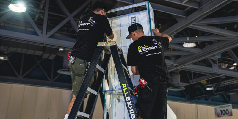 Workers on ladders securing booth walls at a trade show.