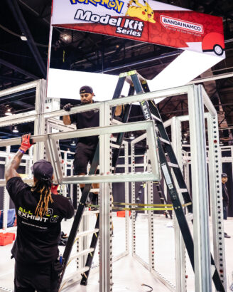 Crew members assembling booth frame with ladders on trade show floor.