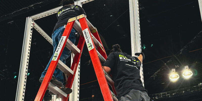 Worker on ladder assembling overhead booth frame at trade show.